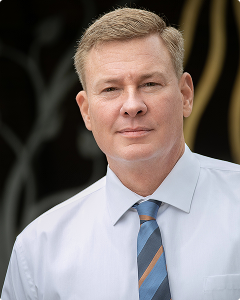 A man with short light hair wearing a light blue dress shirt and a striped tie, standing in front of a dark background with abstract designs.