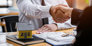 Two people shake hands over a desk with a small model house, documents, and a clipboard, suggesting a real estate or property agreement.