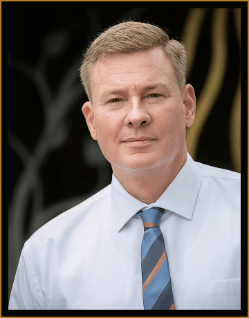 A middle-aged man with short light brown hair wearing a light blue dress shirt and a striped tie poses in front of a blurred dark background.