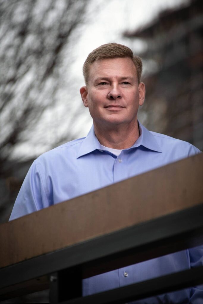 A man in a light blue dress shirt stands outdoors near a railing, with blurred trees and buildings in the background.