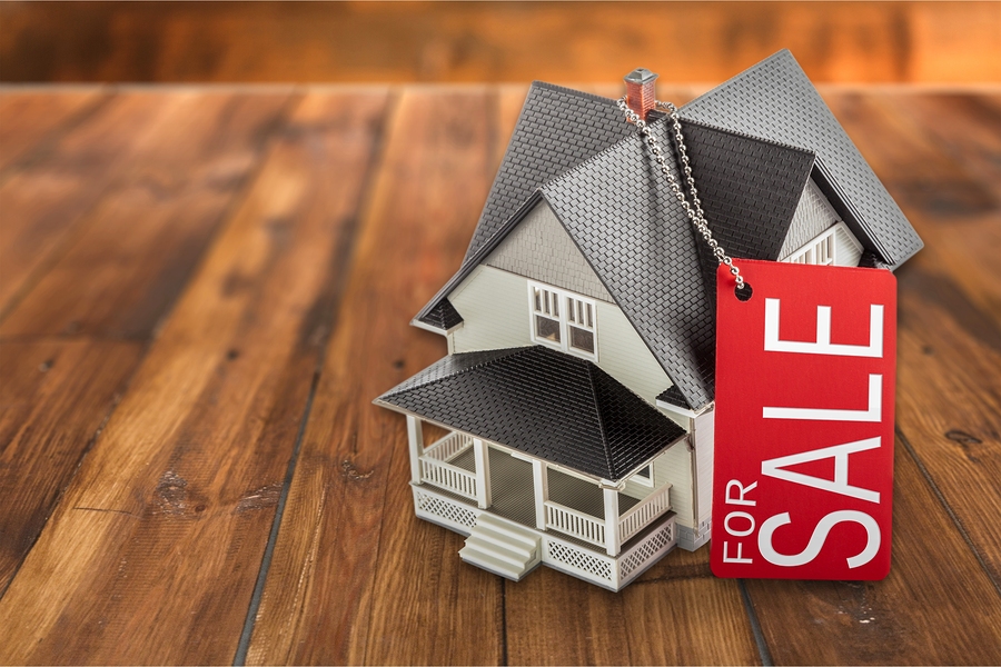 A model house sits on a wooden surface with a large red "For Sale" tag attached to the roof.